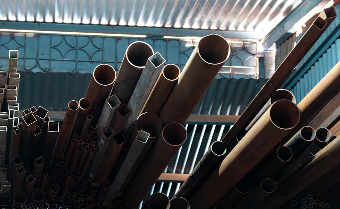 An upward view of an industrial interior, dominated by a dynamic arrangement of metal pipes and square tubes of varying sizes. The contrast between the rusted, circular, and angular forms provides a rich geometric texture. The backdrop of corrugated metal and a bright skylight emphasizes the rugged, raw atmosphere of the storage space. This image is excellent for engineering and construction websites, conceptual design work focused on scale and material, or as a bold, directional background.